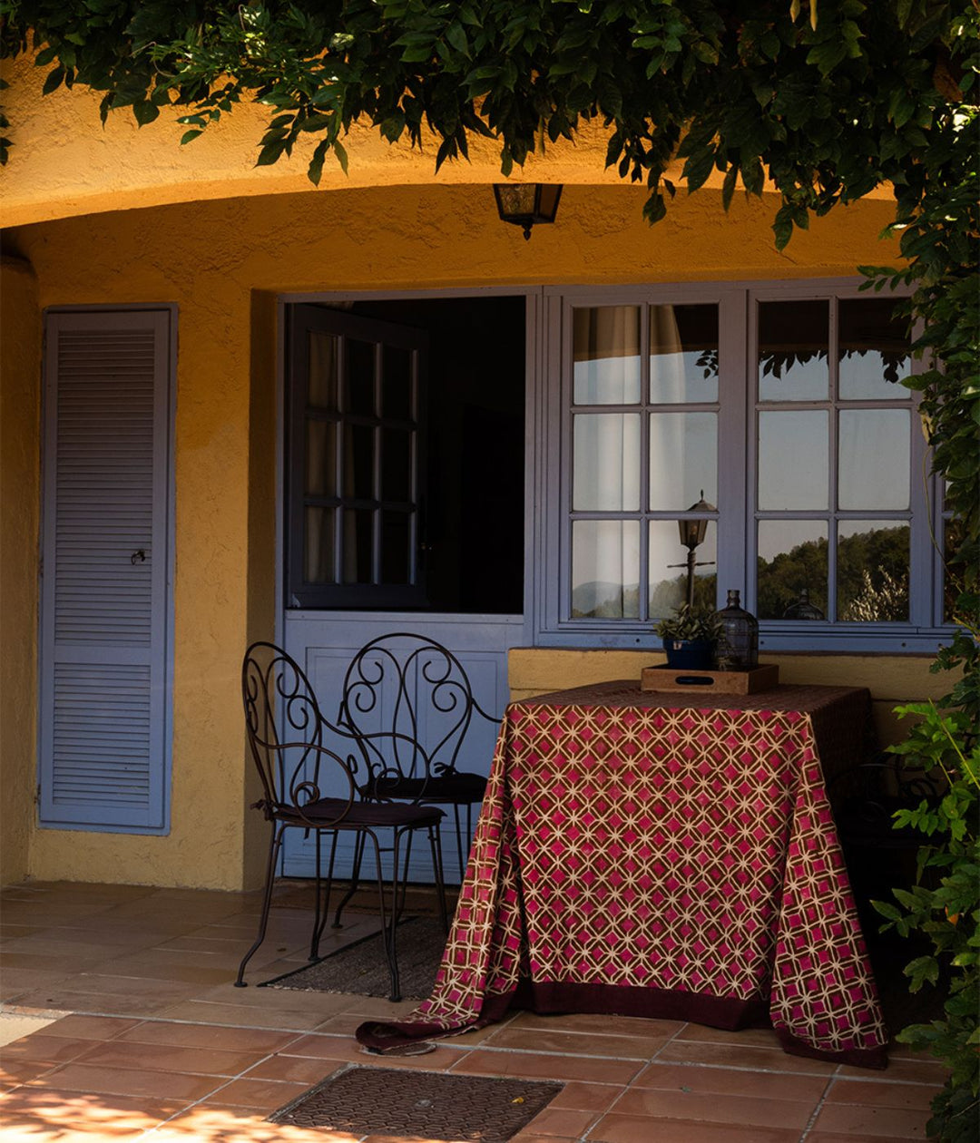 Telo tovaglia artigianale con motivo geometrico rosso e oro su sfondo verde, bordato in tessuto bordeaux, fatto a mano. Utilizzato come tovaglia su un tavolino da due nella veranda di una casa in campagna. 