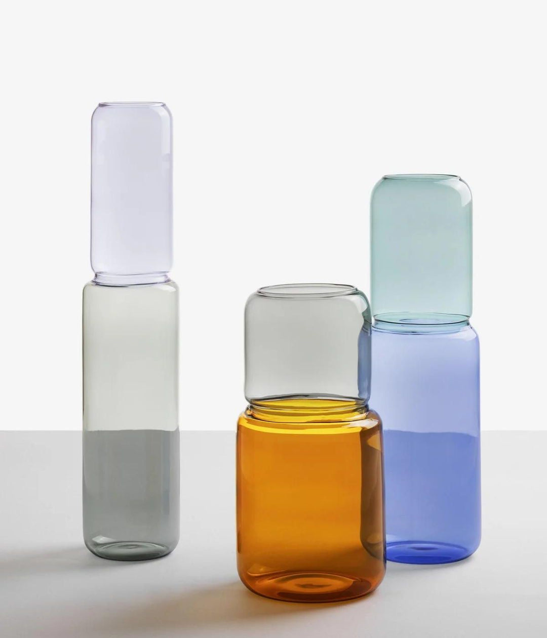 Tre vasi Revolve di Ichendorf in vetro borosilicato, mostrati in colori grigio, ambra e azzurro, disposti su una superficie bianca.