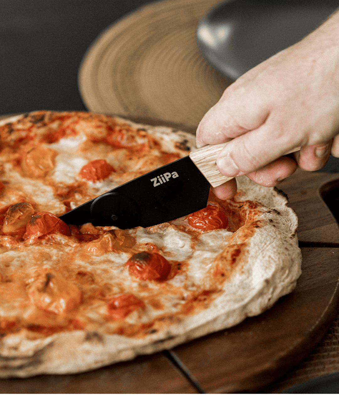 Pizza con pomodorini e coltello con lama nera con rotella che taglia la pizza. Sfondo tagliere marrone.