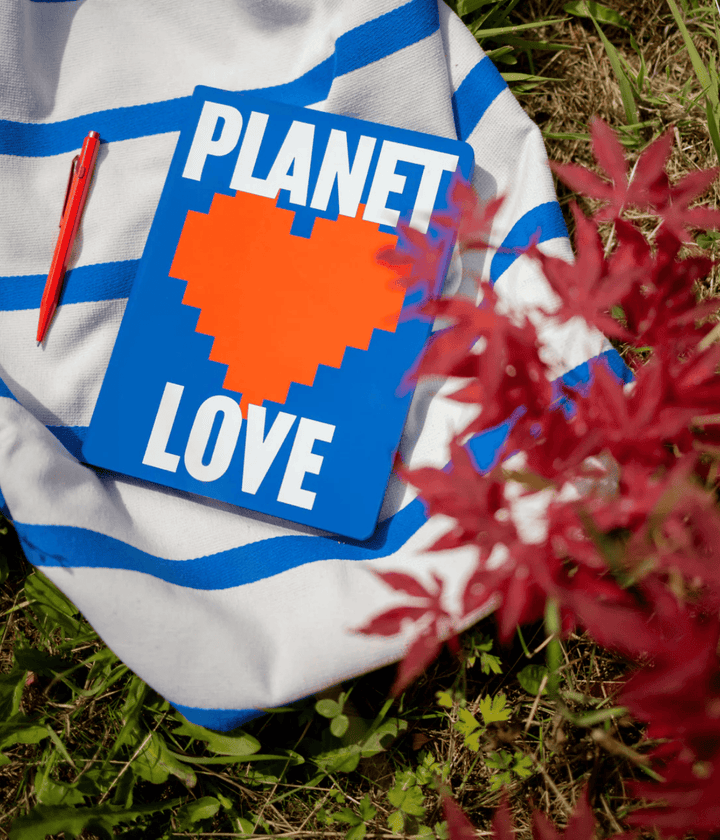 Notebook scritta Planet Love cuore rosso, appoggiato su tela  righe bianche e blu. Sfondo giardino.