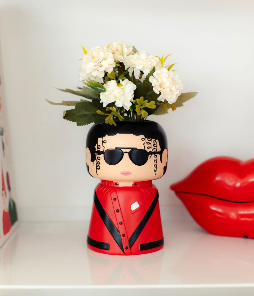 Un vaso in poliresina a forma di Michael Jackson, con occhiali da sole e abbigliamento rosso e nero, utilizzato come vaso per fiori bianchi, posizionato su una mensola accanto a una decorazione a forma di labbra rosse.